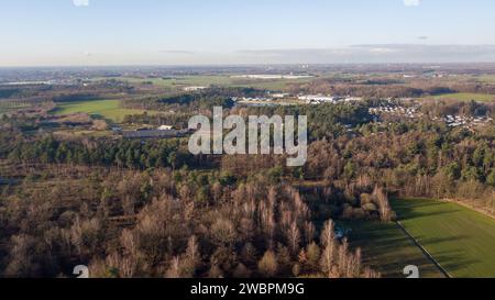 Cette photographie aérienne offre une vue d'ensemble sur un paysage diversifié où les différentes utilisations des terres convergent. Une bande de champs agricoles dans différentes nuances de vert suggère une agriculture active, tandis que des parcelles de bois denses offrent un contraste naturel. La présence de bâtiments industriels à l’horizon indique une activité économique, et un quartier résidentiel se niche à la lisière du milieu naturel, illustrant l’équilibre entre développement et nature. Le ciel ouvert et la vue lointaine vers l'horizon transmettent l'immensité de la zone, avec des nuages projetant des ombres fugaces sur la scène. Vue aérienne Banque D'Images