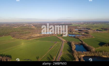 Cette image aérienne capture la beauté expansive de la campagne, caractérisée par des champs verdoyants qui s'étendent au loin, suggérant des terres agricoles fertiles. Une série de petits lacs ou étangs ajoute une touche de bleu au paysage verdoyant, améliorant sa qualité pittoresque. Une route de campagne serpente à travers les champs, offrant la connectivité à travers ce cadre bucolique. En toile de fond, un soupçon de développement urbain à l’horizon offre un contraste subtil avec le premier plan rural. Le ciel clair et la lumière douce d'une journée approchant de sa fin projettent de longues ombres et enrichissent les couleurs de t Banque D'Images