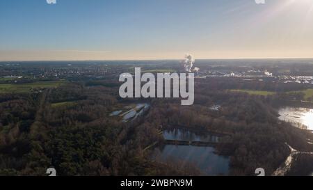 Cette photo aérienne contraste la beauté tranquille d'un paysage naturel avec l'agitation lointaine de l'activité industrielle. Au premier plan, des plans d'eau sereins sont entourés de forêts denses, reflétant le ciel calme et clair. Au fur et à mesure que la vue s’étend, elle capture les champs ouverts, qui servent de tampon avant de céder la place au complexe industriel. Des cheminées ponctuent l'horizon, libérant des panaches dans l'air, un rappel brutal de l'activité économique de la région. La lumière du soleil s'enflamme dans le coin, suggérant que l'image a été capturée tard dans la journée, mettant en évidence l'interaction complexe entre l'induse Banque D'Images