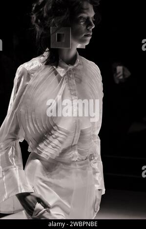 Mannequin portant un chemisier blanc transparent à manches longues avec des rayures blanches avec un grand appliqué rond en tissu superposé et un pantalon blanc au Runway Fashion Show Banque D'Images