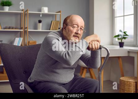 Homme âgé assis à la maison en rééducation avec sa béquille et regardant triste la caméra. Banque D'Images