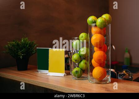 Un grand vase en verre rempli de limes et d'oranges en couches, créant une pièce décorative vibrante sur une surface en bois dans un cadre confortable. Banque D'Images