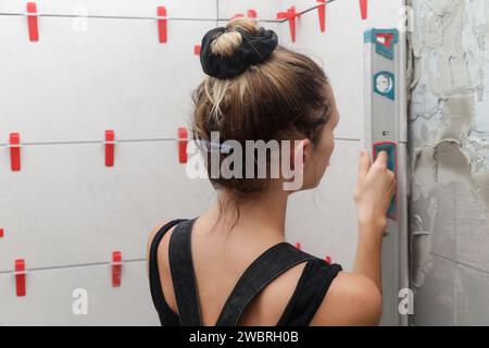 Femme carrelant les murs de salle de bains, en utilisant un niveau à bulle pour mesurer un mur sur le chantier de construction. Processus d'installation de carreaux dans la salle de bain étape par Banque D'Images