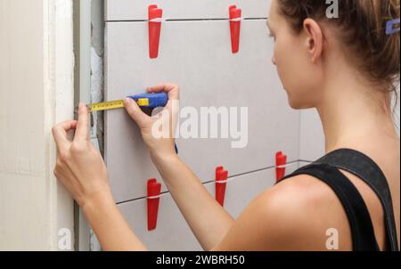 Femme carrelant les murs de salle de bains, en utilisant le mètre ruban pour mesurer la longueur. Processus d'installation de carreaux dans la salle de bain étape par étape. Bricolage maison Improvem Banque D'Images