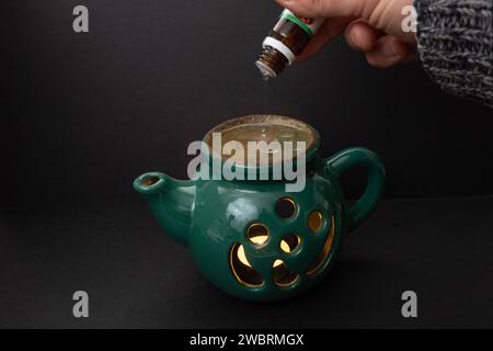 Bougie brûleur à huile essentielle en forme de théière verte, avec une main versant une petite bouteille sur fond sombre, Banque D'Images