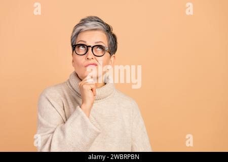 Photo de grand-mère intelligente esprit toucher le menton look intéressé espace vide isolé sur fond de couleur beige Banque D'Images
