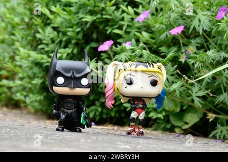 Funko Pop figurines d'action de DC comics super-héros Batman et Harley Quinn debout sur la route asphaltée dans le parc de printemps, herbe verte, fleurs roses. Banque D'Images