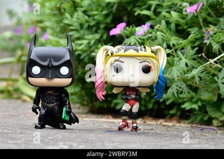 Funko Pop figurines d'action de DC comics super-héros Batman et Harley Quinn debout sur la route asphaltée dans le parc de printemps, herbe verte, fleurs roses. Banque D'Images