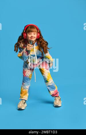 Petite fille heureuse avec le syndrome de Down portant des écouteurs, le microphone holing et chantant sur fond de studio bleu. Passe-temps Banque D'Images