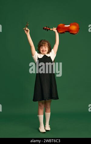 Belle fille avec le syndrome de Down debout avec violon, apprenant la musique sur fond de studio vert Banque D'Images