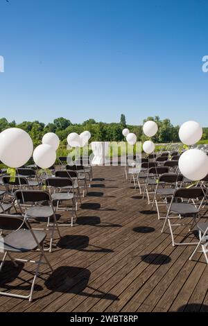 Cette image capture l'installation tranquille d'une cérémonie en plein air au bord d'un lac, avant l'arrivée des invités. La scène est composée de rangées de chaises métalliques soigneusement disposées sur une terrasse en bois, chacune ornée d’un ballon blanc, ajoutant une touche d’élégance festive. Les ballons captent la lumière et se balancent doucement dans la brise, créant une atmosphère ludique mais sereine. Au point focal de l'arrangement se dresse un podium solitaire, drapé de tissu blanc, attendant l'arrivée de l'orateur ou du couple. Le ciel bleu clair et la verdure luxuriante en arrière-plan complètent le cadre pittoresque de ce qui promet d'être Banque D'Images