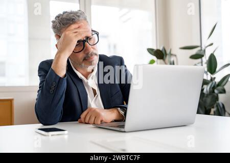 Employé de bureau masculin réfléchi utilisant un ordinateur portable à l'intérieur, regardant l'écran d'ordinateur portable avec l'expression de visage perplexe, homme d'affaires résolvant des tâches, ayant des problèmes avec les finances Banque D'Images