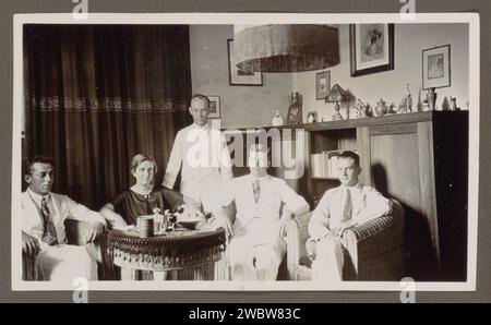 Quatre hommes et une femme autour d'une table, anonyme, 1920 - 1930 photographie trois hommes en vêtements blancs et une femme sont dans des fauteuils en osier dans une pièce, un quatrième homme se tient entre eux. Sur la table ronde se trouvent des verres avec des boissons, une bouteille et un bol. Le capuchon en tissu d'une lampe suspendue est toujours visible. Sur la droite contre le mur se trouve un placard avec quelques livres. Sur elle comprend une statue d'un chat, une photo encadrée, un lampadaire et des vases. Sur les murs accrochent des images encadrées, y compris une nature morte. Fait partie de l'album familial Dutch East Indies. support photographique impression argentique gélatine intérieur de la maison Banque D'Images