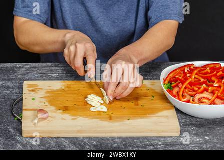 Mains de femme hachant l'ail frais sur une planche de bois. Saladier sur table grise, fond noir Banque D'Images