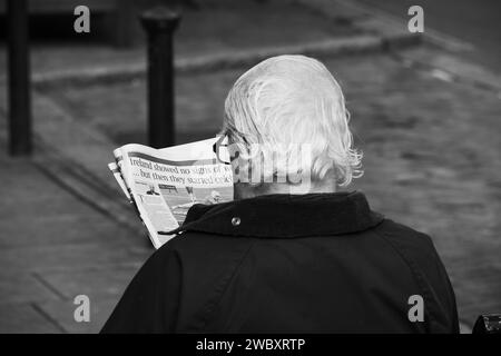 Ross on Wye, Herefordshire, Angleterre septembre 24 2023 : un homme aux cheveux gris et aux lunettes s'assit à lire un journal sur un banc public. Photo noir et blanc Banque D'Images