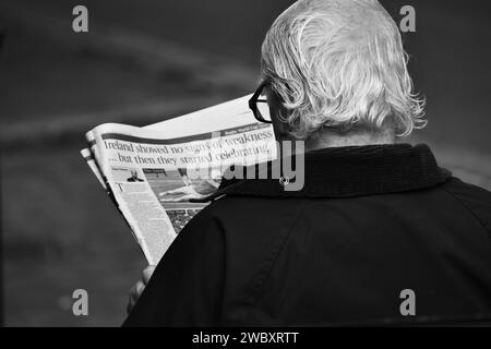 Ross on Wye, Herefordshire, Angleterre septembre 24 2023 : un homme aux cheveux gris et aux lunettes s'assit à lire un journal sur un banc public. Photo noir et blanc Banque D'Images