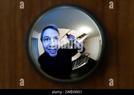 vue à travers le spectateur de porte dans un escalier d'un grand immeuble, peephole, avec un homme en face de la porte souriant et tenant une bouteille de vin rouge Banque D'Images