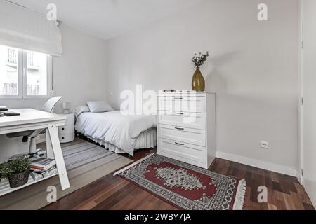 Chambre meublée avec un lit simple, un bureau en bois blanc, divers tapis au sol et une commode assortie Banque D'Images