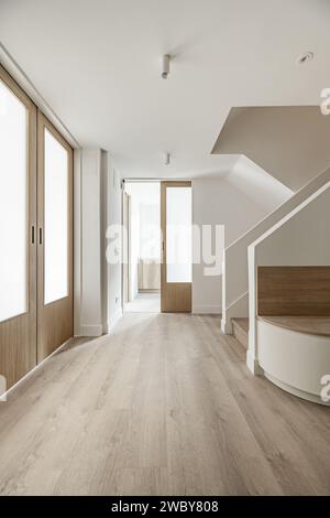 Hall d'entrée d'une maison avec des murs peints en blanc lisse, armoires encastrées blanches, escalier, parquet assorti et portes coulissantes en bois et en verre Banque D'Images