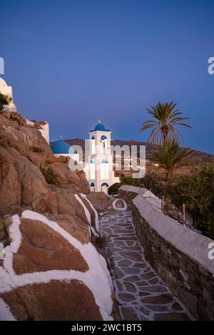 Un chemin menant à l'église orthodoxe blanchie à la chaux illuminée de Panagia Gremniotissa à iOS Grèce, entourée de palmiers au coucher du soleil Banque D'Images