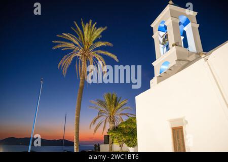 Vue de l'église orthodoxe blanchie à la chaux illuminée de Panagia Gremniotissa à iOS Grèce, entourée de palmiers au coucher du soleil Banque D'Images