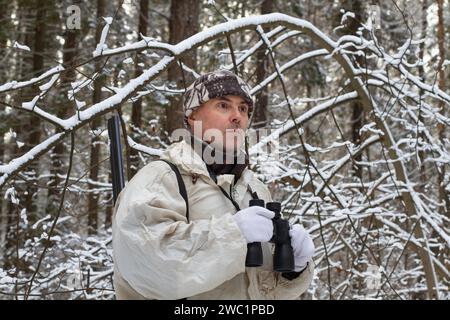 Un chasseur en camouflage blanc se tient dans la forêt nordique enneigée. Il tient les jumelles dans ses mains. Gros plan. Banque D'Images