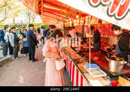 Femme japonaise tentant dans son sac à main pour acheter un poulpe à un stand de nourriture takoyaki dans le parc du château d'Osaka pendant le festival des cerisiers en fleurs. Banque D'Images