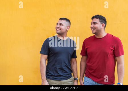 Engagés et heureux, deux hommes en chemises colorées regardent vers le haut avec des sourires, debout devant une toile de fond jaune frappante. Banque D'Images