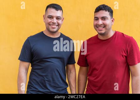 Une paire d'hommes, l'un en chemise sombre et l'autre en rouge, partagent un sourire amical sur fond jaune vif, rayonnant une camaraderie détendue. Banque D'Images