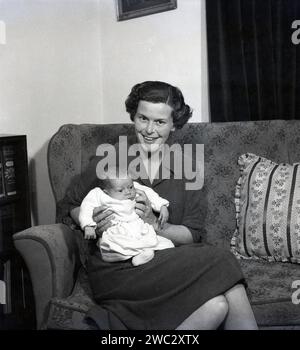 Années 1950, historique, une dame dans une robe en laine assise sur un canapé avec un nouveau-né sur ses genoux, le montrant à la caméra, Angleterre, Royaume-Uni. Dans la bibliothèque du coin, le livre relié, The Scientific Romances of H. G. Wells, publié pour la première fois au début des années 1930 Banque D'Images