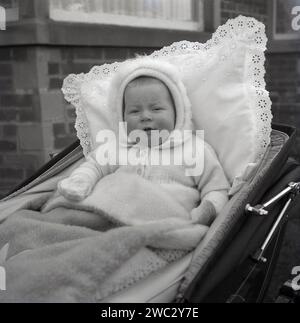 années 1960, historique, extérieur, enveloppé, la tête couverte, gants et cardigan sur et sous une couverture de laine et avec un coussin recouvert de dentelle confortable derrière lui, un enfant en bas âge assis dans un landau construit par autocar de l'époque, Angleterre, Royaume-Uni. Banque D'Images