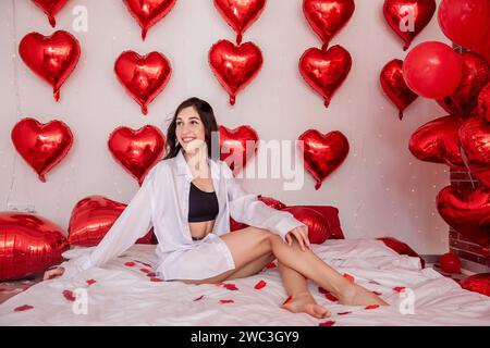 Belle femme assise sur le lit orné de draps blancs, souriant. Fille blogueuse entourée de ballons en forme de coeur rouge attachés au mur, scattere Banque D'Images