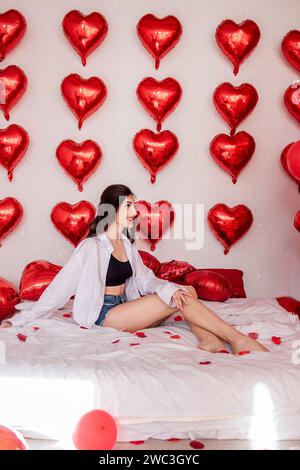 Belle femme assise sur le lit orné de draps blancs, souriant. Fille blogueuse entourée de ballons en forme de coeur rouge attachés au mur, scattere Banque D'Images