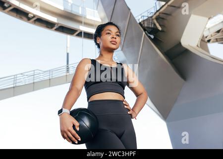 Jeune femme mince avec les cheveux bouclés en tenue de fitness noire tenant un ballon de médecine debout à l'extérieur Banque D'Images