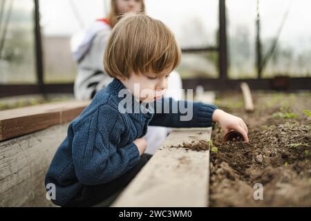 Mignon garçon en bas âge jouant avec la terre le jour du printemps. Kid plantant un semis dans une serre. Enfant explorant la nature. Activités estivales pour les petits enfants. Banque D'Images