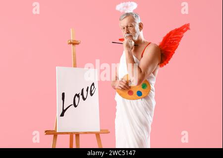 Homme mûr réfléchi habillé comme Cupidon Holding peintures et chevalet avec mot AMOUR sur fond rose. Fête de la Saint-Valentin Banque D'Images