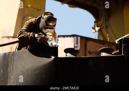 Soudeur qualifié dans l'équipement de protection au travail, joignant des pièces métalliques sur l'équipement industriel. Des étincelles jaillissent de la torche de soudage en usine. Banque D'Images