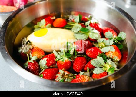 Fraises rouge vif et feuilles vertes flottant dans l'eau dans un bol en acier inoxydable, se préparant à une friandise rafraîchissante ou à une utilisation culinaire. Banque D'Images