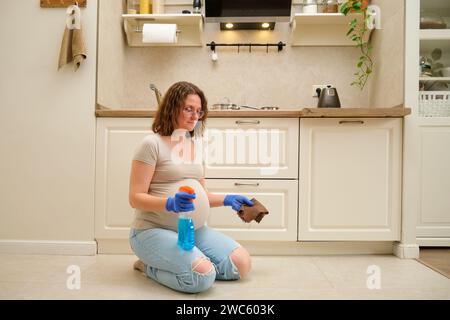 La femme enceinte fait des travaux ménagers dans la cuisine, en utilisant un chiffon pour essuyer le sol. Banque D'Images