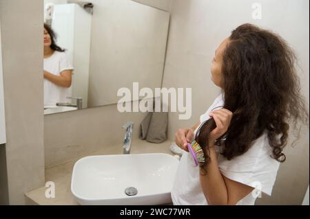 Vue arrière d'une jeune jolie femme adulte avec de longs cheveux bruns bouclés, peignant les cheveux avec une brosse à cheveux, souriant regardant son reflet dans le bathroo de la maison Banque D'Images