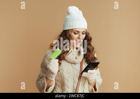 Bonjour l'hiver. femme élégante excitée en pull beige, mitaines et chapeau sur fond beige avec smartphone et carte de crédit achetant sur internet. Banque D'Images