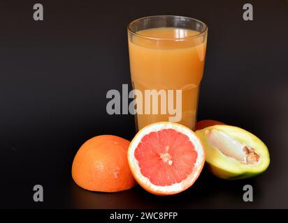 Un mélange de jus de fruits dans un grand verre sur fond noir, à côté de morceaux de pamplemousse mûr et de mangue. Gros plan. Banque D'Images