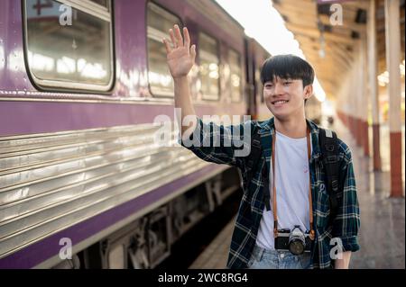 Un jeune homme asiatique heureux et beau en vêtements décontractés agite la main, disant bonjour à quelqu'un à une gare. Personnes et concepts de transport Banque D'Images