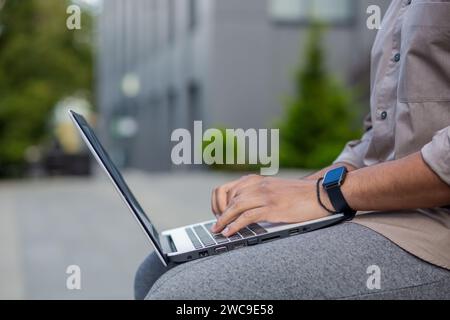Gros plan d'une personne utilisant un ordinateur portable à l'extérieur, concentrez-vous sur les mains avec une smartwatch, symbolisant le travail à distance, la technologie et la mobilité. Banque D'Images