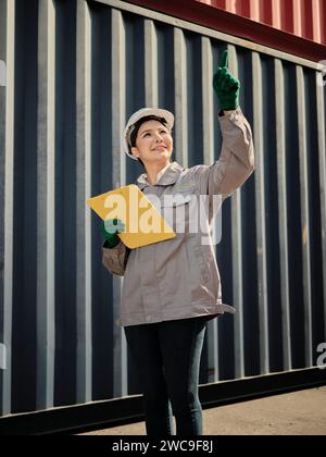 Portrait de femme asiatique travailleur tenant le presse-papiers et pointant avec l'index sur le fond du conteneur d'expédition Banque D'Images