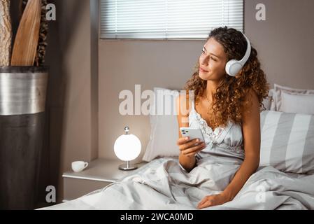 Jeune femme calme portant des vêtements de nuit assis dans le lit à l'aide d'un smartphone tout en écoutant de la musique sur des écouteurs de la playlist dans la chambre à coucher Banque D'Images