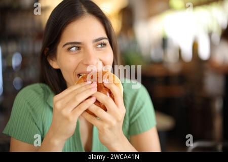 Femme heureuse mangeant un hamburger regardant de côté dans un intérieur de restaurant Banque D'Images