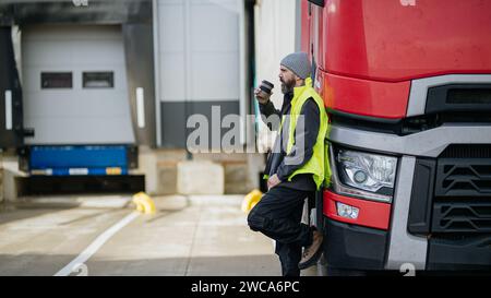 Chauffeur de camion appuyé contre le camion rouge et boire du café, attendant les travailleurs de l'entrepôt. Banque D'Images