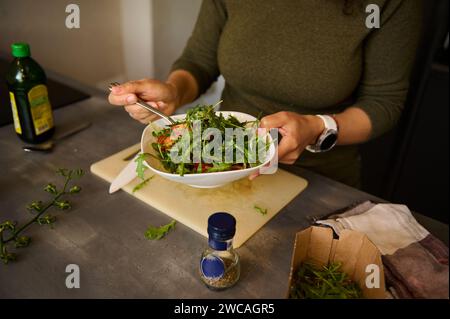 Gros plan des mains féminines tenant un bol blanc de salade fraîche saine avec des légumes verts, roquette et tomates. Vue recadrée d'une jeune femme cuisinant sainement Banque D'Images