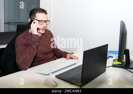 Un secrétaire homme tient une conversation sur smartphone sur son bureau devant un ordinateur portable Banque D'Images